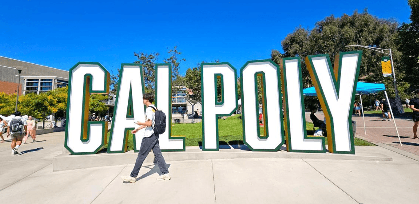 Large white and green letters spelling CAL POLY on a campus walkway with people walking nearby.