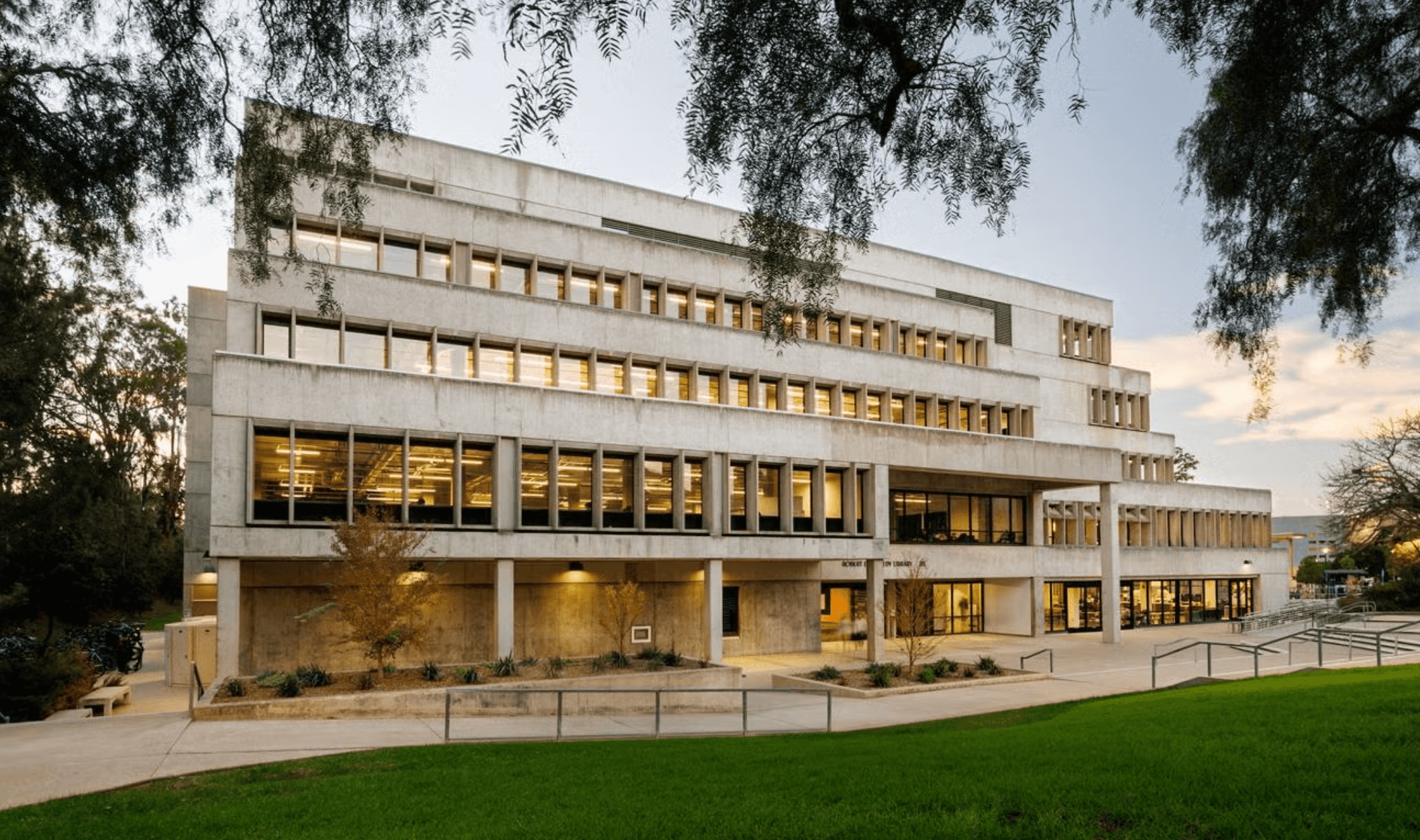 Modern concrete multi-story building with large windows, surrounded by trees and green lawn at dusk.