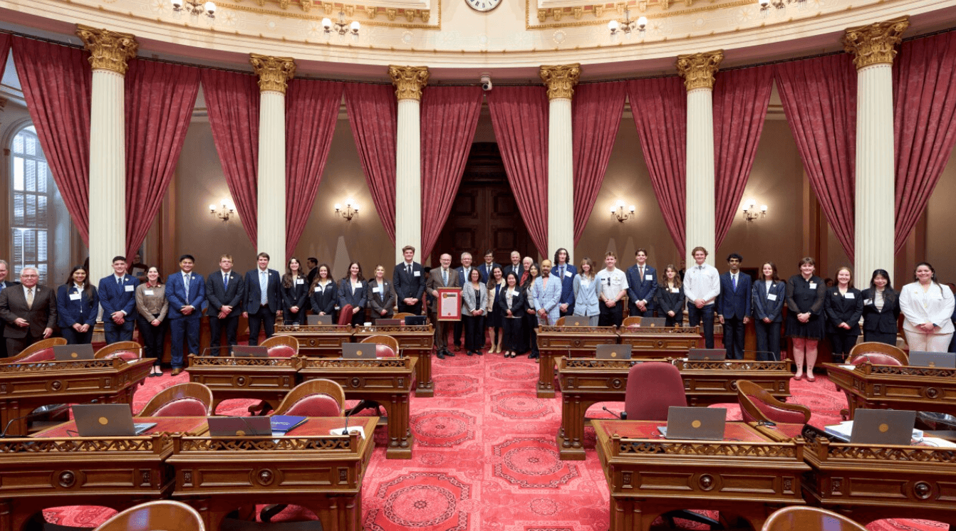 Group of people in formal attire standing in a room with red curtains, gold-trimmed columns, and wooden desks with laptops.