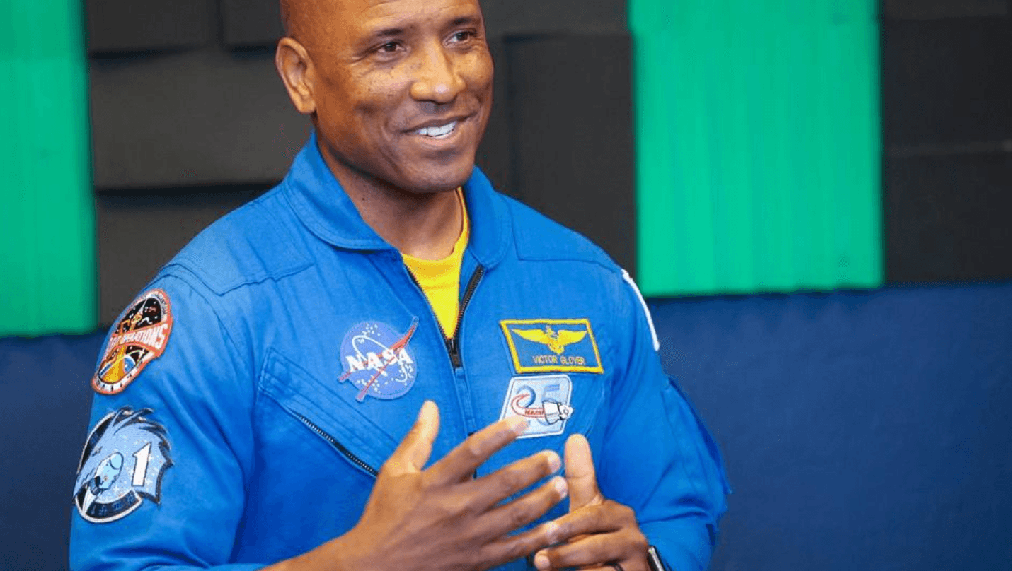 Man in a blue NASA flight suit with patches speaking, hands gesturing.