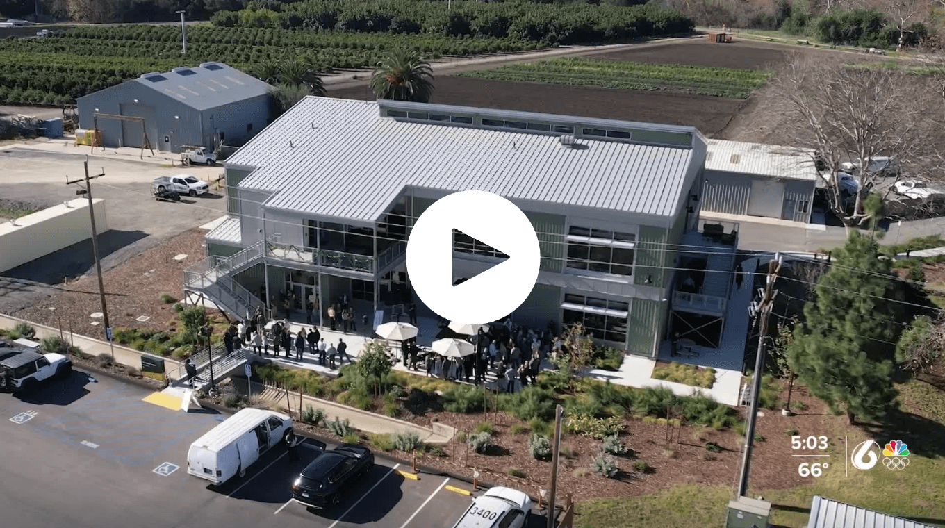 Crowd gathered outside a modern two-story building with metal roof and large windows on a sunny day.