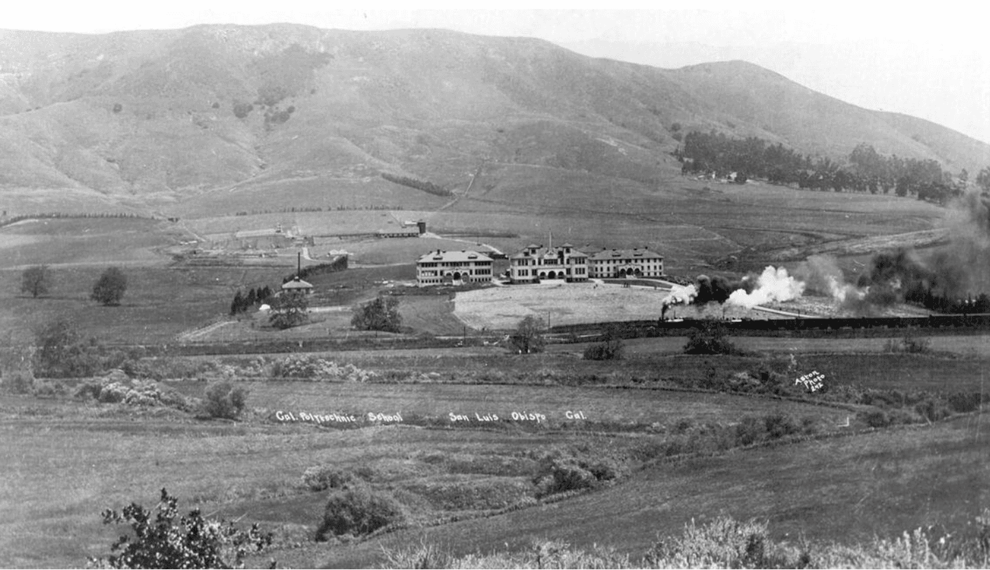 Large campus building in a valley with hills in the background, a steam train passing by leaving smoke trails.