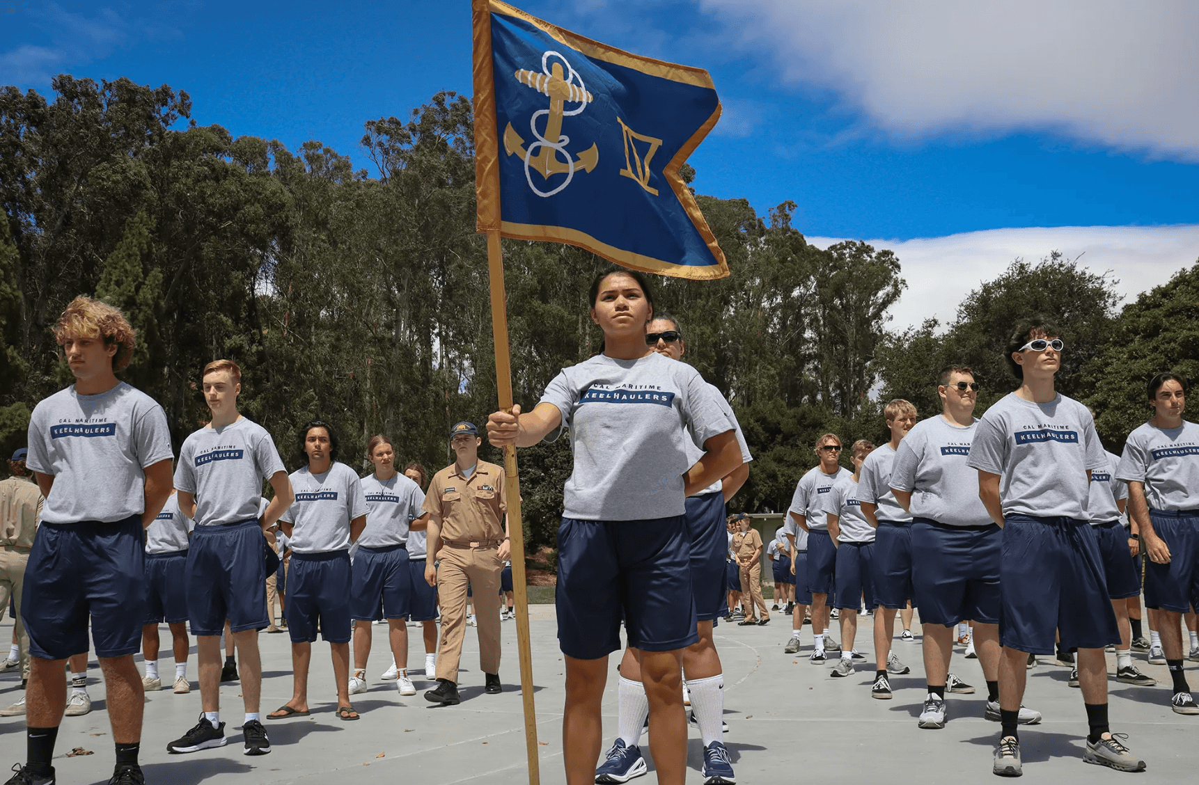 Group of young people in Cal Maritime Keelhaulers shirts standing outdoors with one holding a blue flag with an anchor symbol.