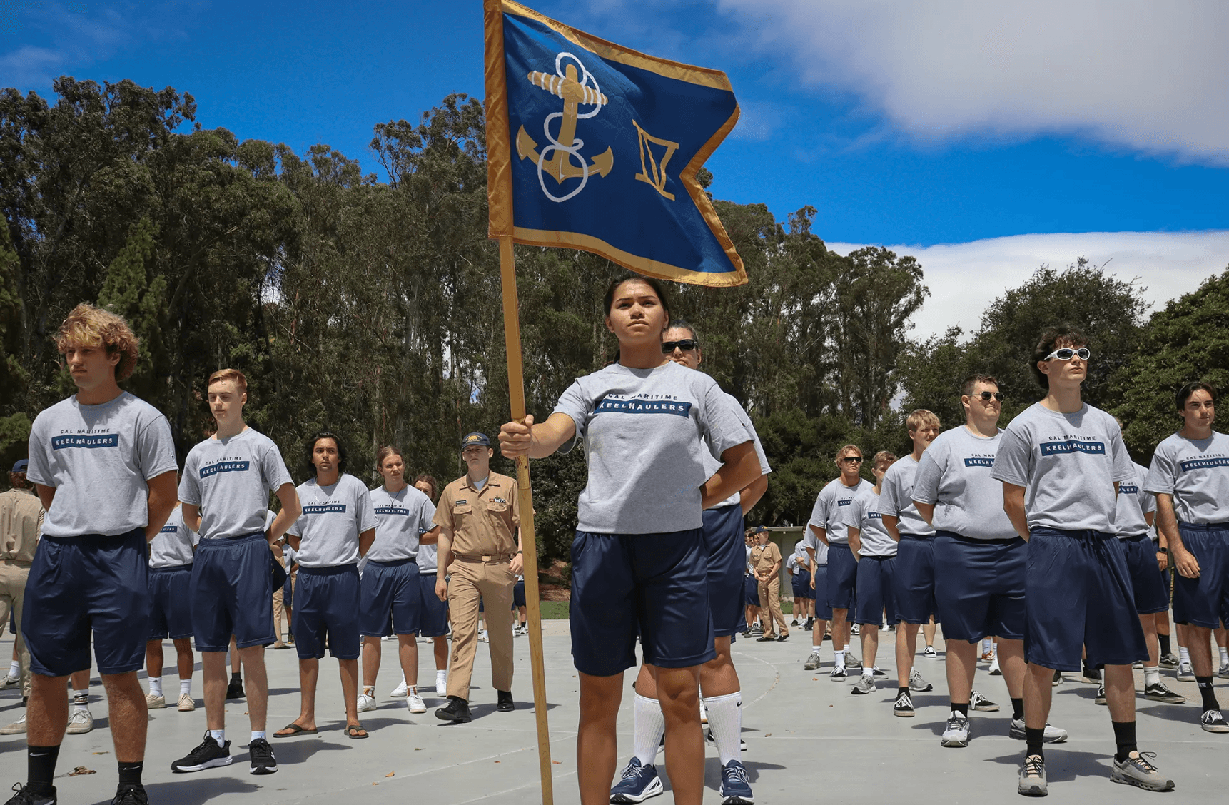 Group of young people in Cal Maritime Keelhaulers shirts standing outdoors with one holding a blue flag with an anchor symbol.