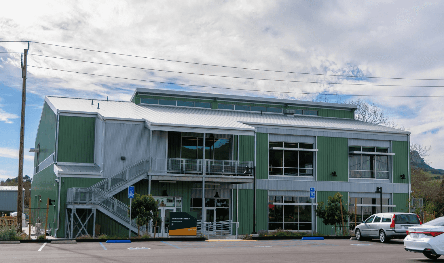 Green and silver two-story building with exterior staircase, large windows, handicap parking, and a sign reading "Technology Park II.