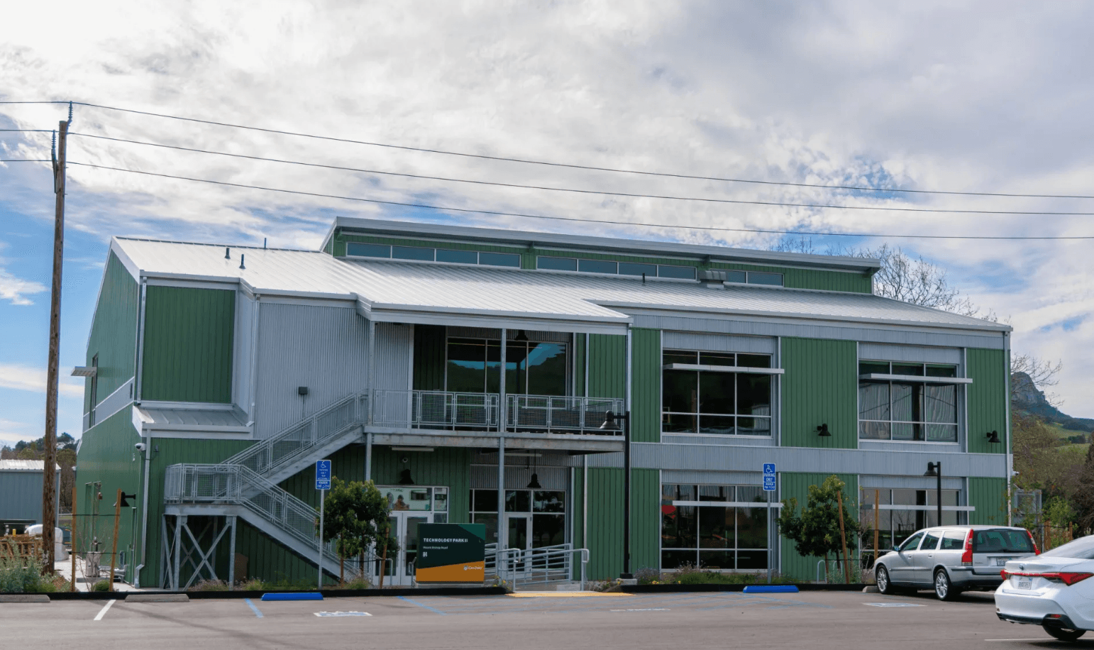 Green and silver two-story building with exterior staircase, large windows, handicap parking, and a sign reading "Technology Park II.