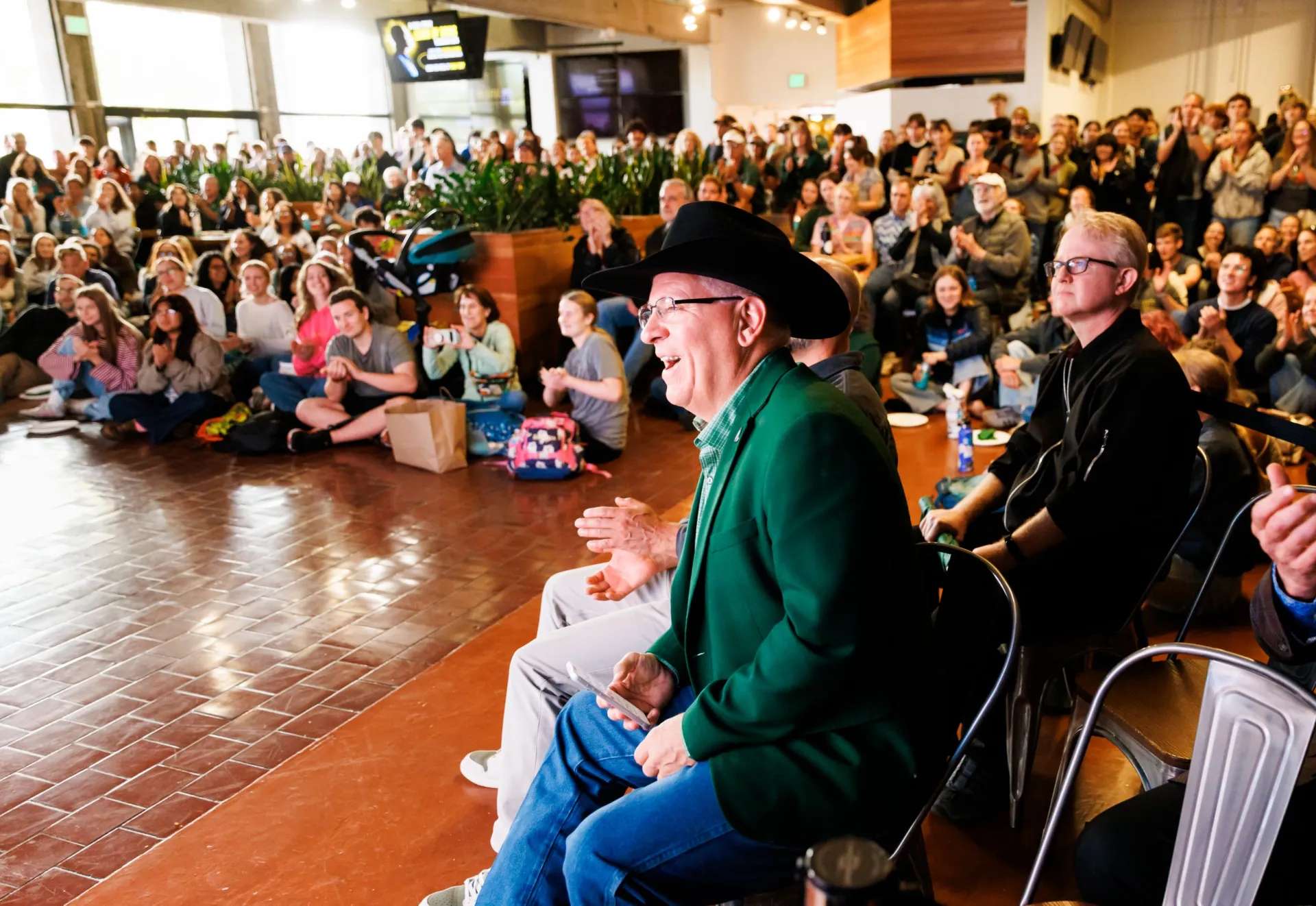 Man in green jacket and black cowboy hat smiling and clapping at an indoor event with seated and standing audience.