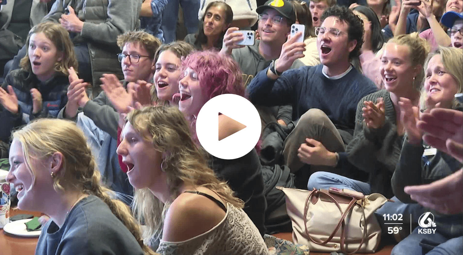 Crowd of people sitting, clapping, and cheering with some recording on phones at an indoor event.