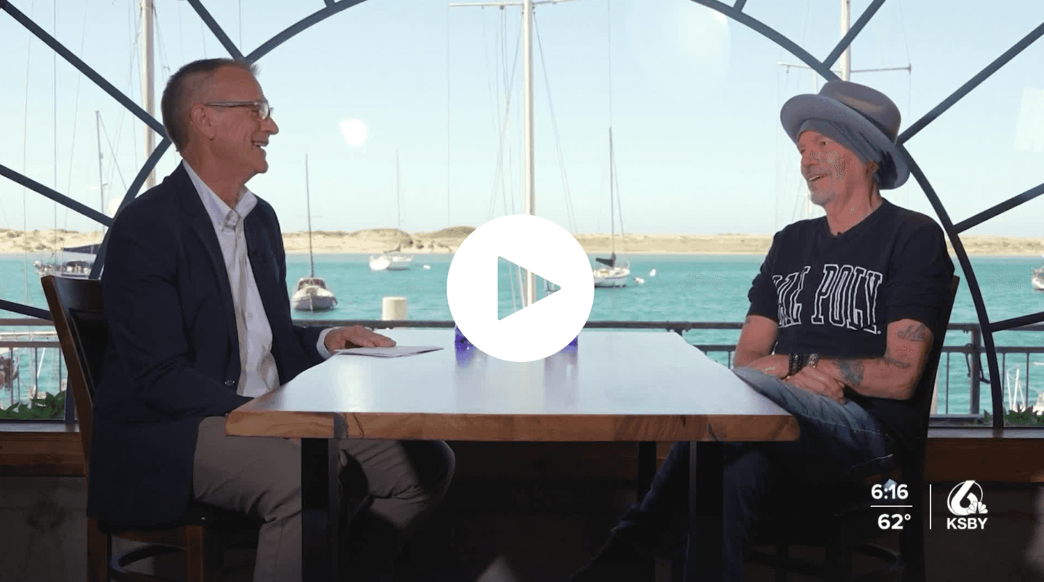 Two men sitting across from each other at a table by a waterfront with boats in the background, KSBY logo.