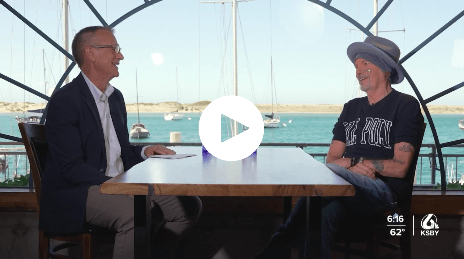 Two men sitting across from each other at a table by a waterfront with boats in the background, KSBY logo.